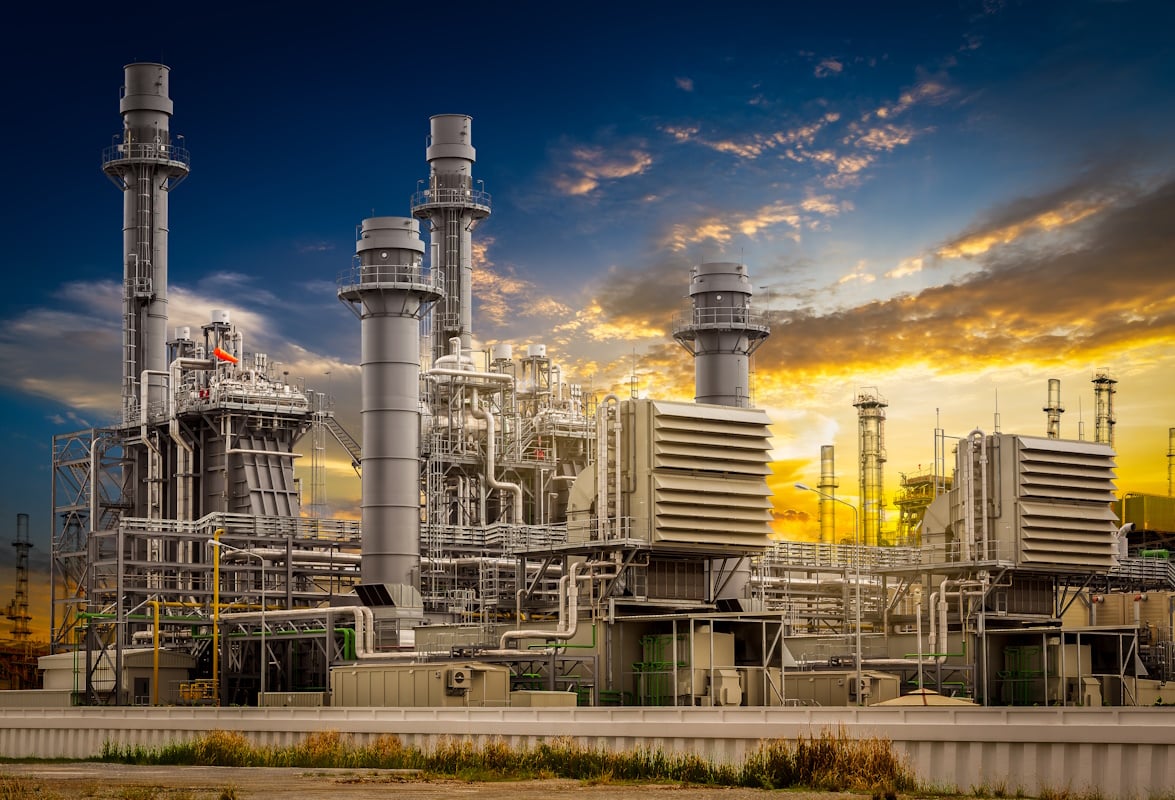 Large power plant complex with towering chimneys and intricate networks of pipes and turbines, set against a dramatic sunset sky.