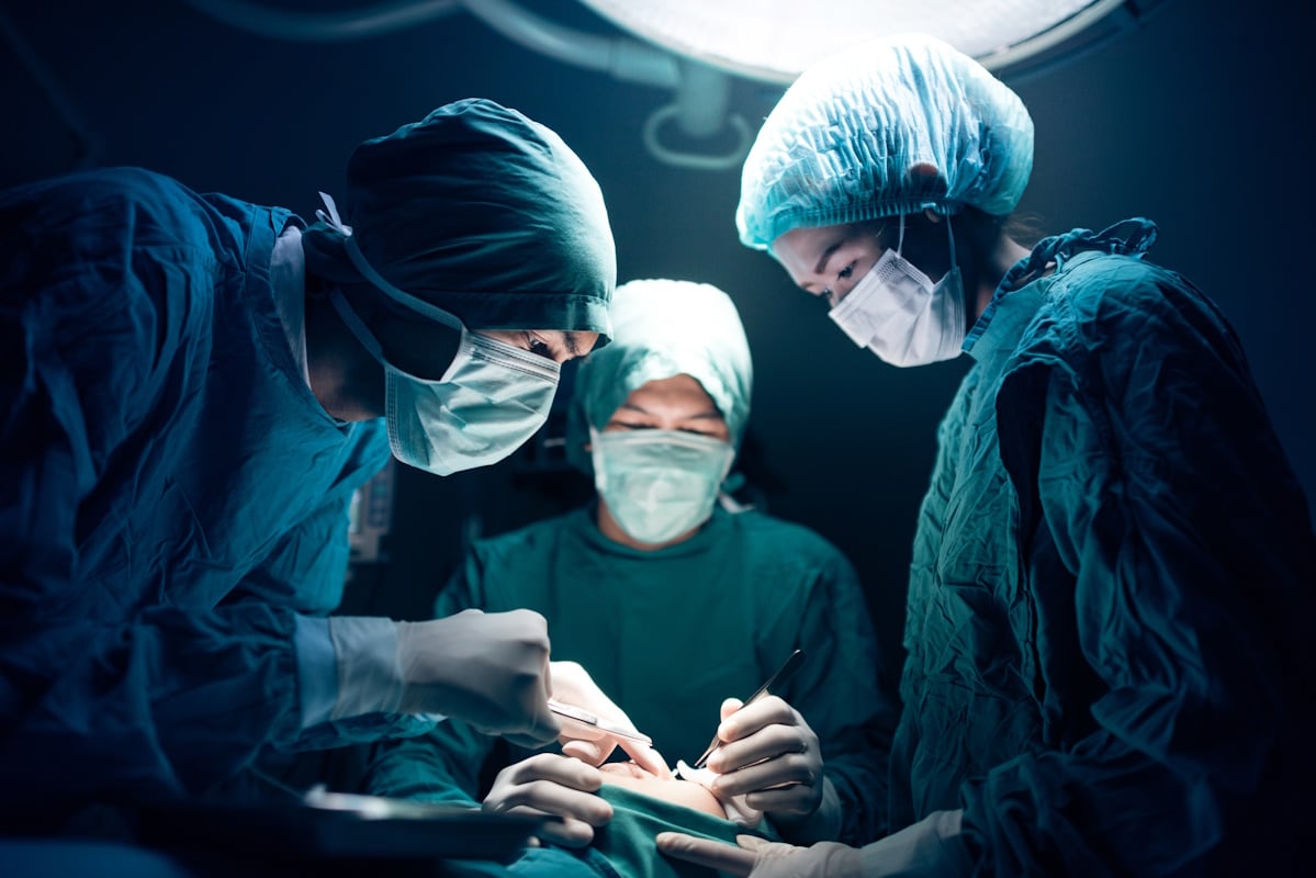 Team of surgeons performing an operation under intense surgical lighting in a sterile operating theatre, all dressed in blue scrubs and masks.